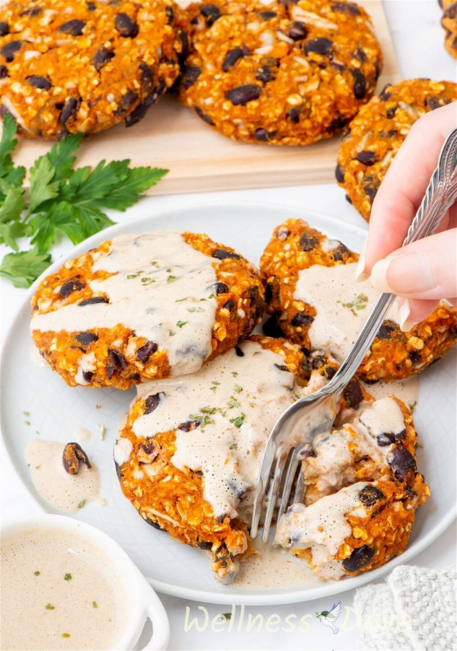 the Sweet Potato Black Bean Burgers in a plate, covered in sauce and a hand is taking a bite with a fork.
