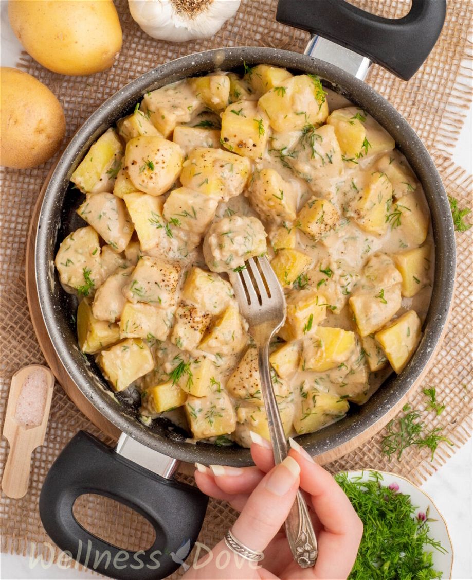 the Garlic Potatoes in a skillet while a hand is taking a potato with a fork - overhead photon