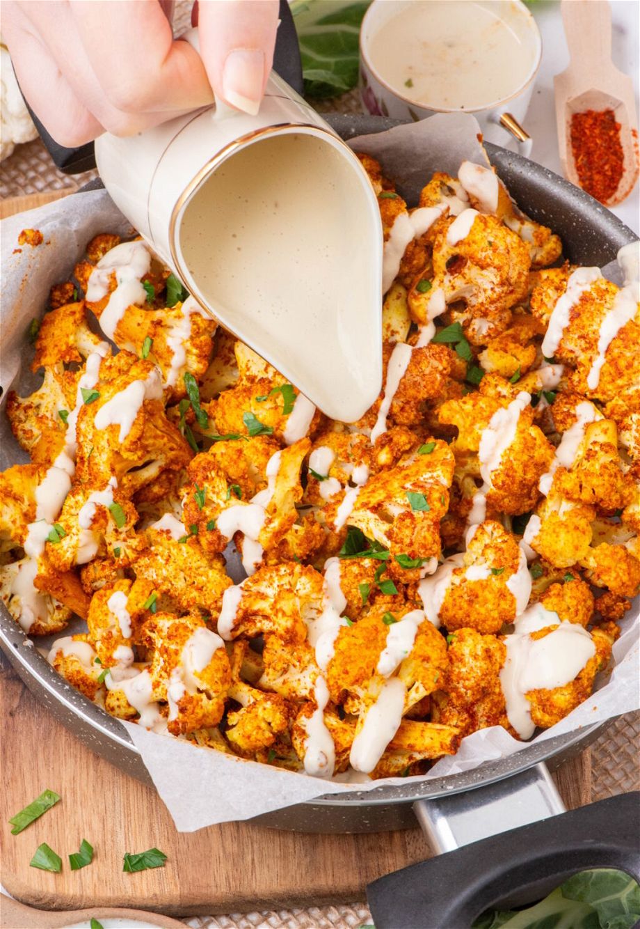 the Oil-free Oven Roasted Cauliflower in a pan and a hand is pouring sauce on top of them.