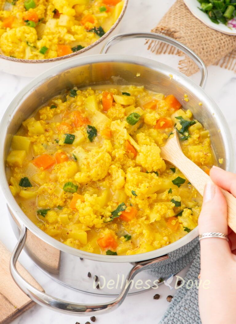 the Cauliflower Vegan Soup with Quinoa in a pot and a hand is stirring it with a wooden spoon